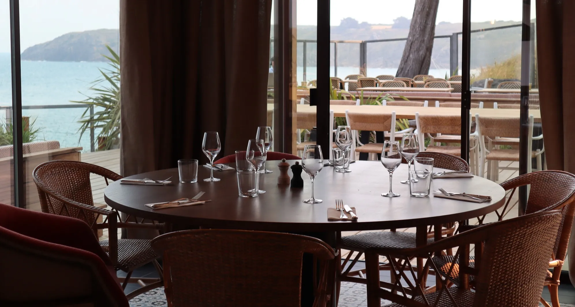 Grande table ronde dressée devant la baie vitrée ouverte sur la mer